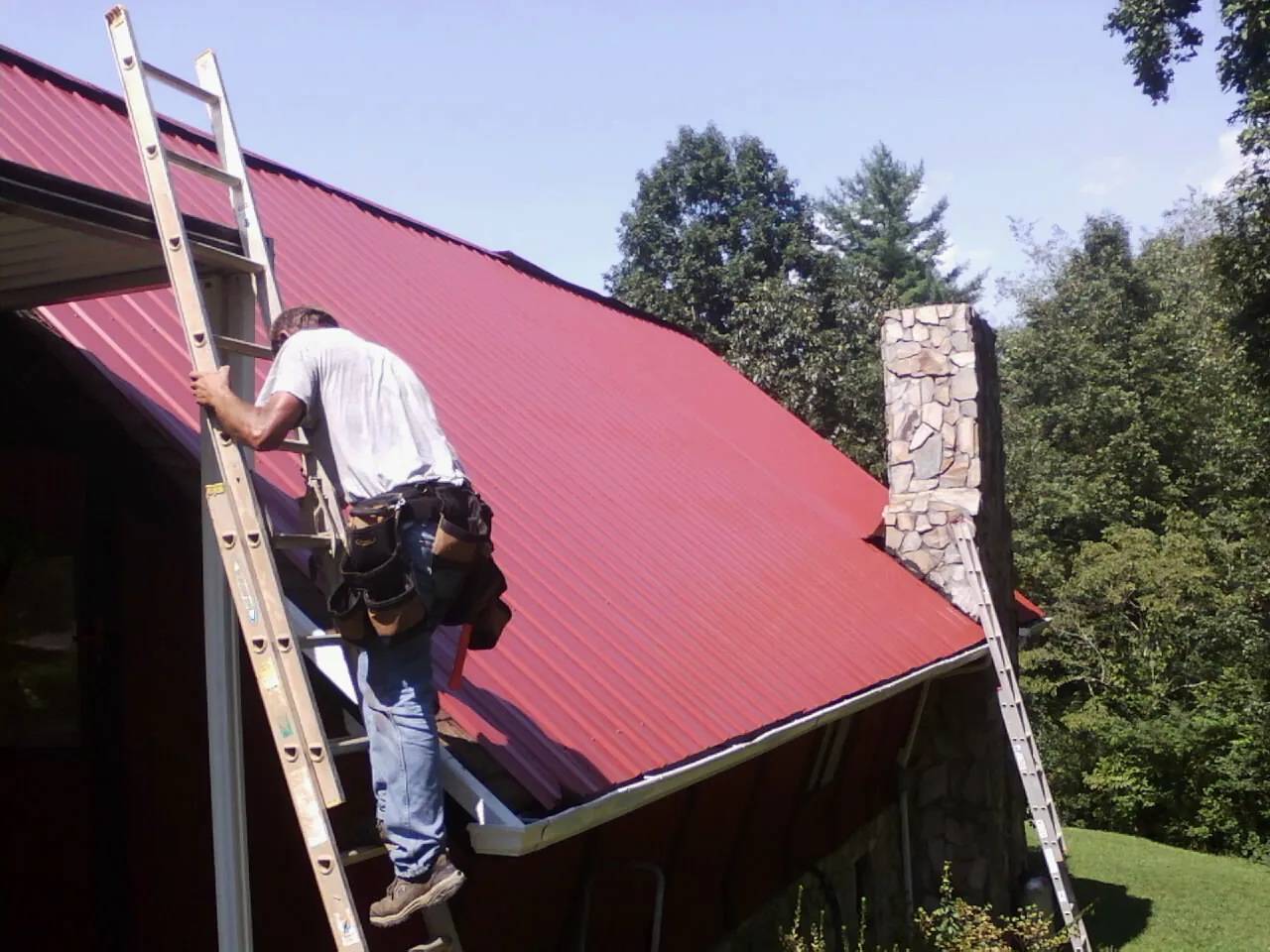 Roofer on a ladder beside a red metal roof and stone chimney — Soffit & Fascia Repair by Roofing Brothers in Biglerville